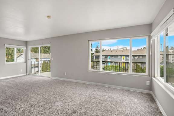 A spacious room with grey carpeting and large windows overlooking a residential area.